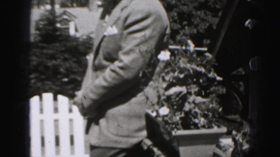 The man appears in partial side view in the 1946 New Cumberland home movie, standing near shrubs, a railing, and a white picket fence.