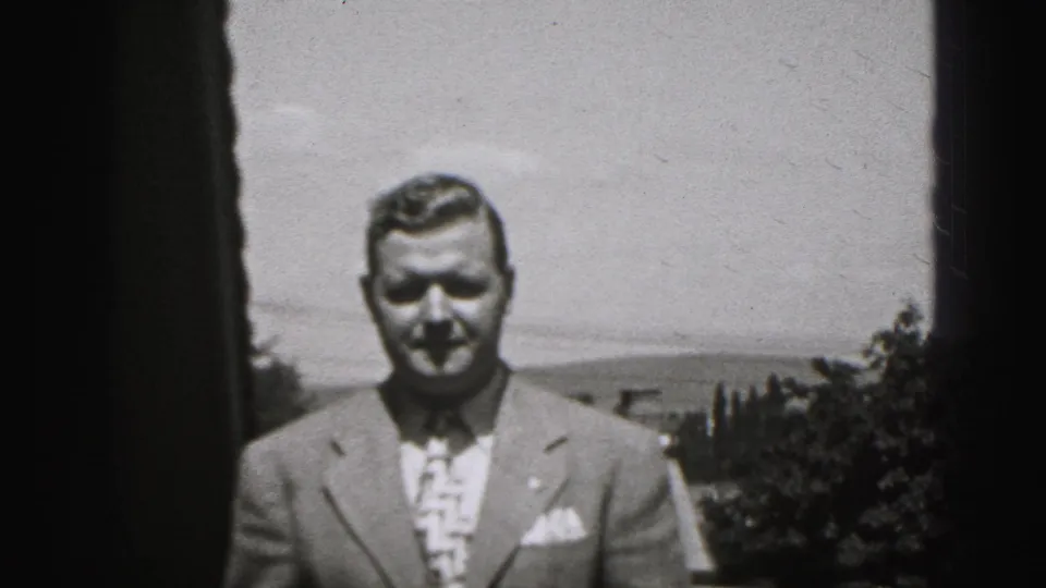 The suited man faces the camera in the 1946 New Cumberland footage, with trees, rooftops, and open sky visible behind him.