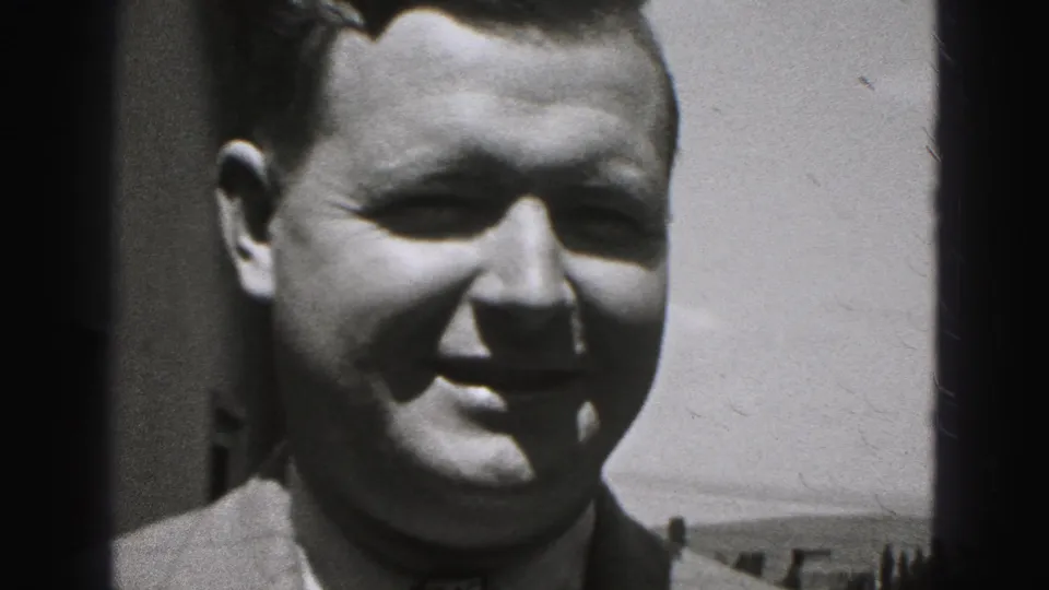 Close-up of the smiling man in suit and tie, with a dark vertical edge and bright outdoor background behind him.