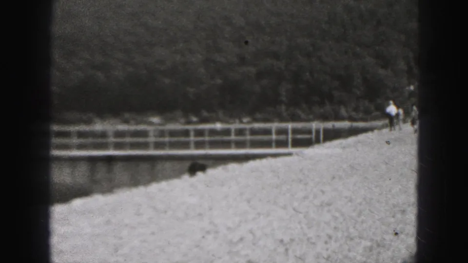 1946 New Cumberland home-movie frame showing a gravel levee path, railing, small dog, and distant pedestrians against a wooded slope.