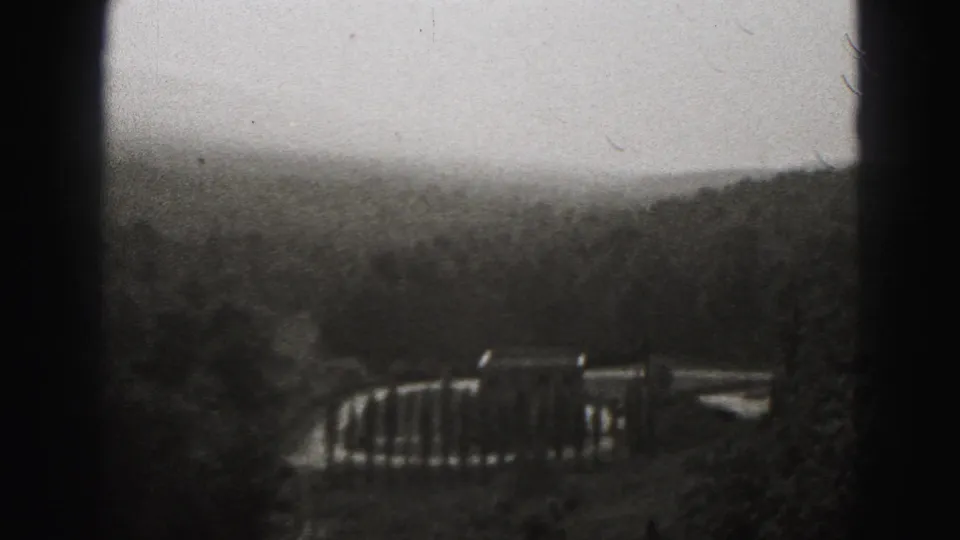 Final 1946 New Cumberland frame looking past a railing toward wooded hills under a pale sky in soft black-and-white 8mm texture.