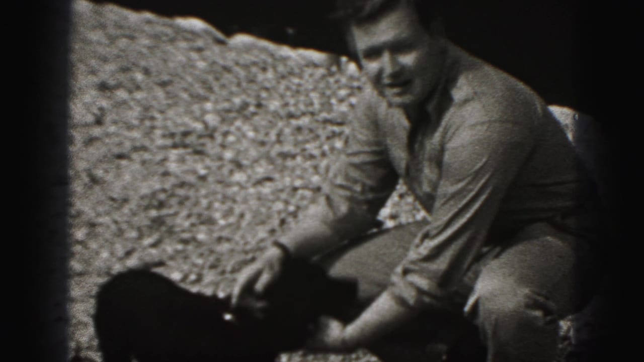Man in a button-up shirt crouches on gravel to pet a small black puppy in 1946 New Cumberland home-movie footage.