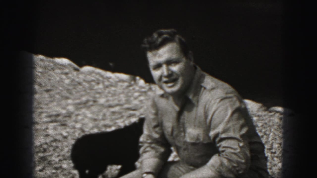 Man crouching on gravel beside a small dark dog in bright sunlight.