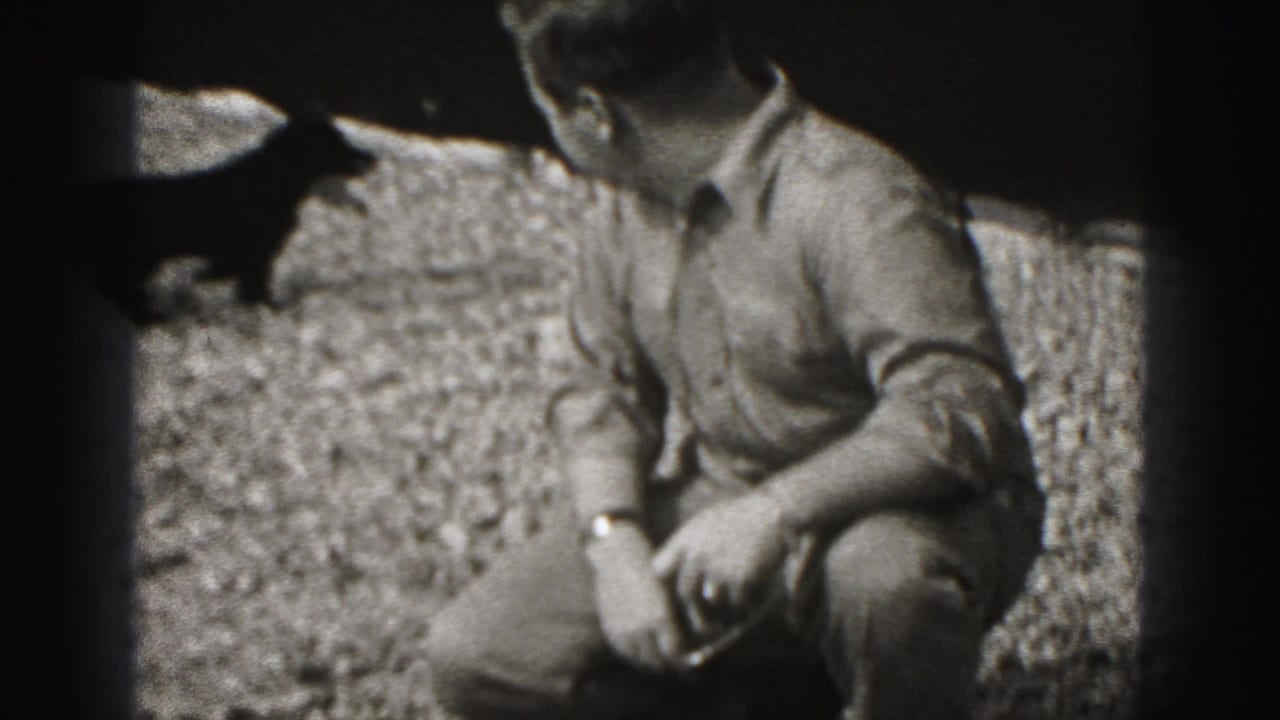 A small dark dog jumps near the crouching man on the gravel, with the man's face partly out of frame in the 1946 home movie.