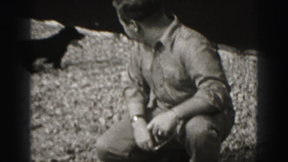 A small dark dog jumps near the crouching man on the gravel, with the man's face partly out of frame in the 1946 home movie.
