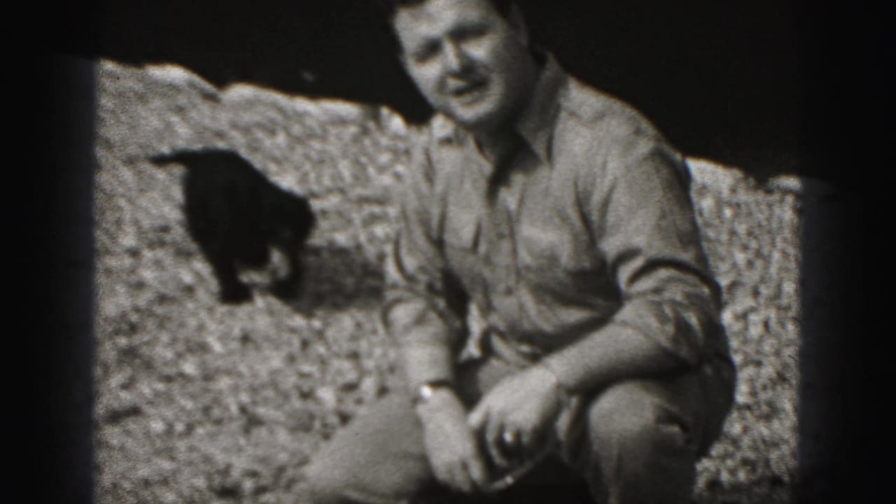 Crouching man smiles beside a small dark dog on gravel.