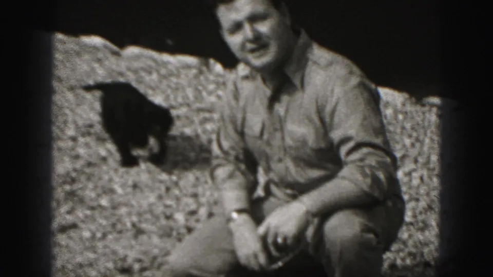 In 1946 New Cumberland home-movie footage, a crouching man smiles near a small dark dog against a rough stone backdrop.