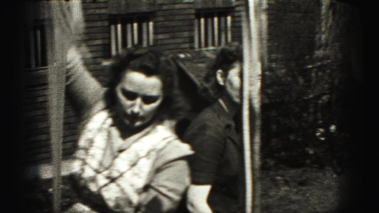 A woman holding a baby stands near a building, with another adult partly visible behind her.