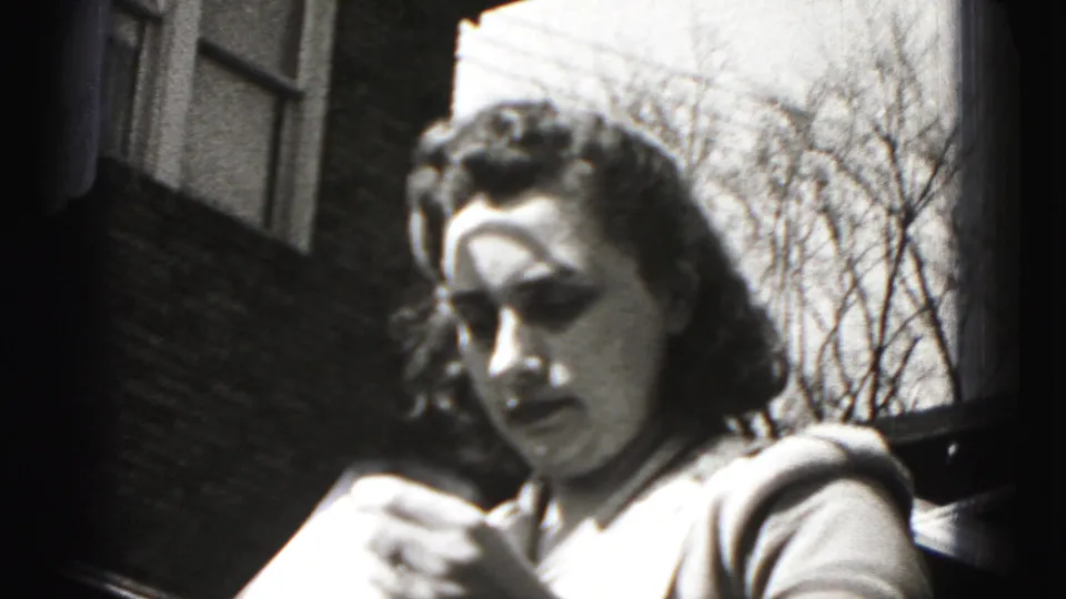 In 1946 New Cumberland, Pennsylvania, a woman in a patterned apron handles fabric near a brick building in bright sun.