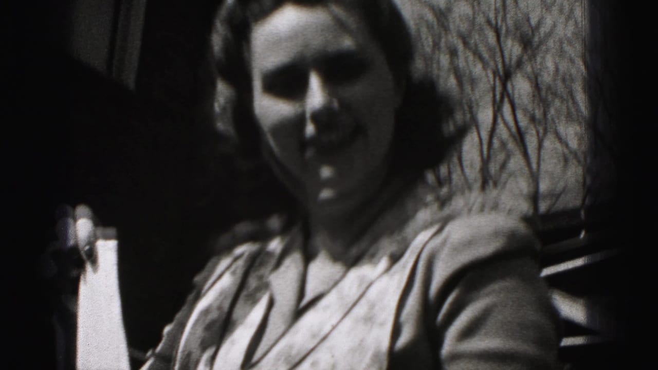 Close handheld 8mm view of the apron-wearing woman outdoors, with her face partly shadowed and bare trees in the background.