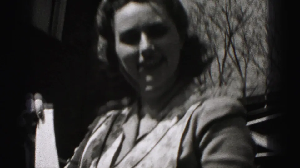 Close handheld 8mm view of the apron-wearing woman outdoors, with her face partly shadowed and bare trees in the background.