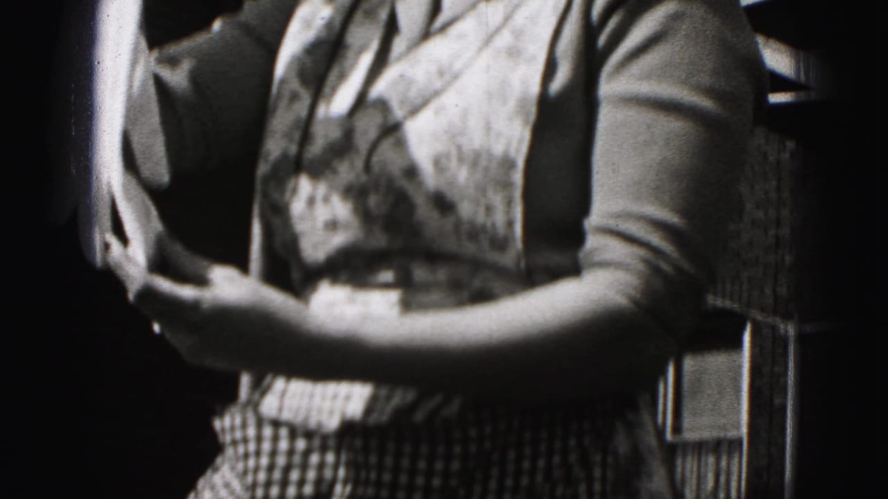 The woman holds fabric in both hands near a brick building, with her checkered lower garment or apron visible in the 1946 footage.