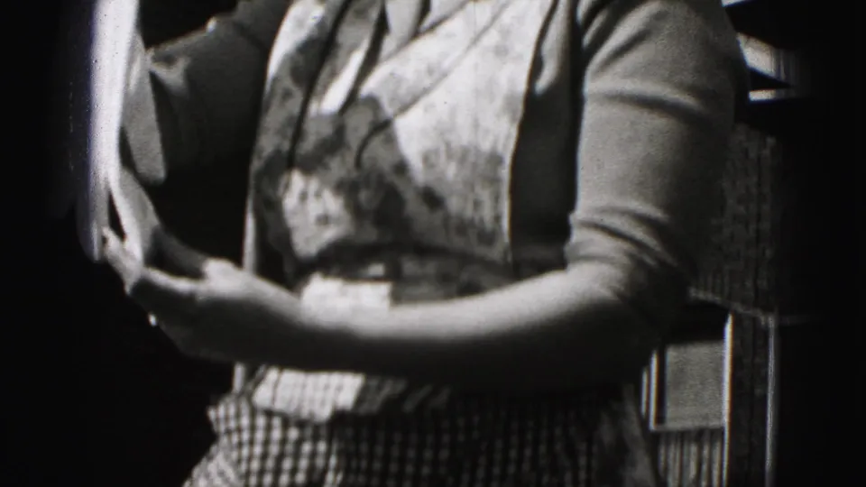 The woman holds fabric in both hands near a brick building, with her checkered lower garment or apron visible in the 1946 footage.