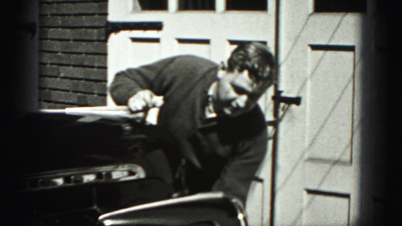 Man bends over the front of a dark vintage car near garage doors.