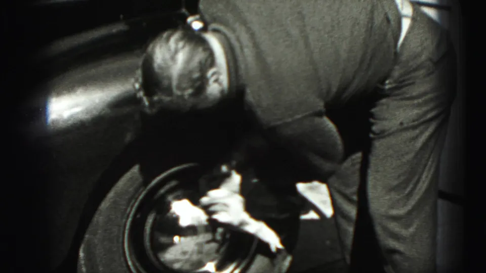The man bends toward the wheel and lower fender of the vintage car, appearing to wipe or inspect the surface.
