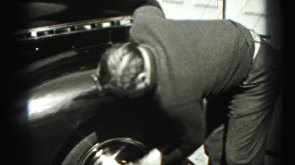 A close view shows the man leaning across the dark fender and wheel area of the car in the 1946 home-movie sequence.