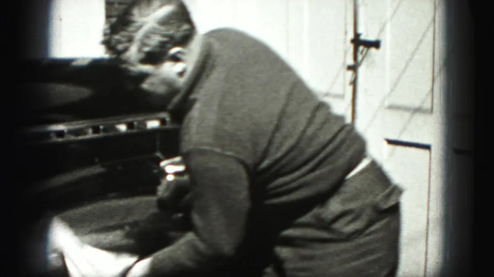 In 1946 New Cumberland, Pennsylvania, a man bends over the front of a dark car while wiping it beside a garage door.