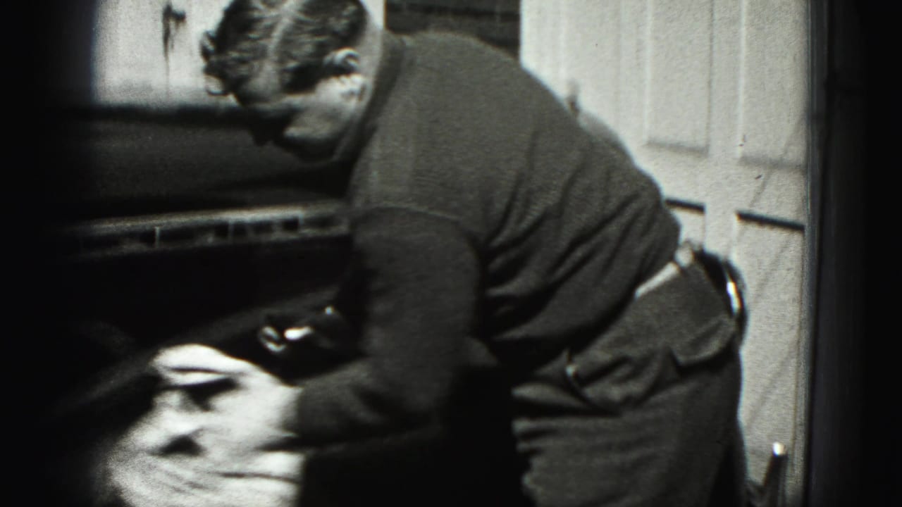 Man wiping the side or front of a dark car beside a paneled garage door.