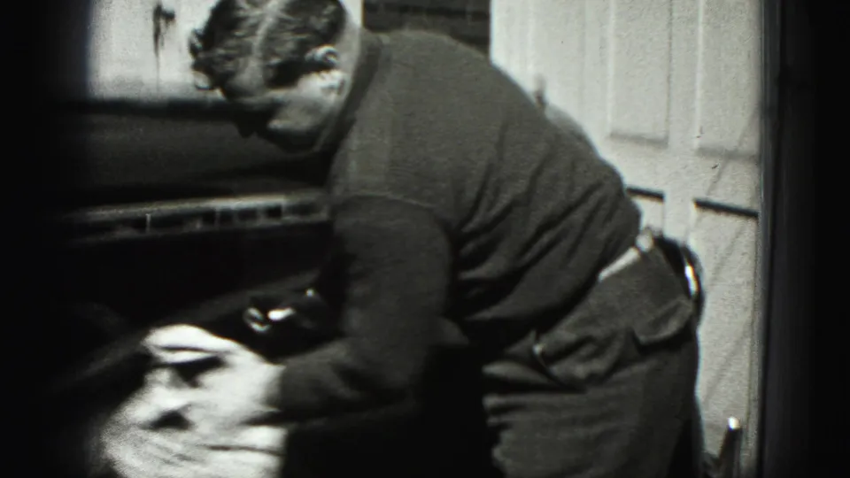 The man continues wiping the automobile in front of a paneled garage door in the New Cumberland home-movie scene.