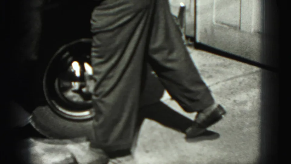 A 1940s home-movie frame shows the man beside the car's wheel, bent forward as he wipes the lower bodywork.