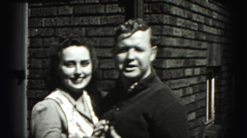An adult couple in New Cumberland, Pennsylvania, smiles close together beside a brick wall in this 1946 8mm frame.