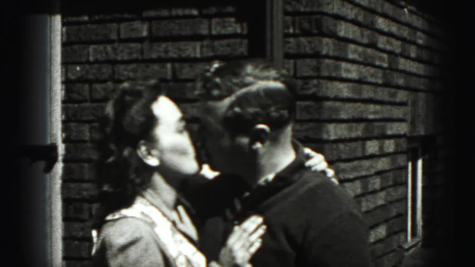 The adult couple kisses in profile beside a brick wall, matching the intimate home-movie setting described for 1946 New Cumberland.