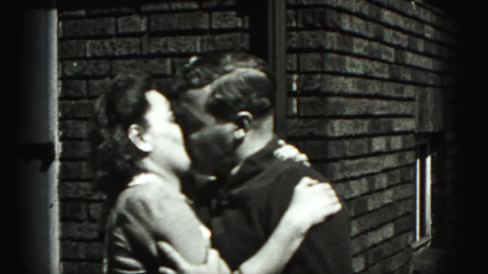 In 1946 New Cumberland home-movie footage, a woman in a light dress kisses a man in a dark sweater by a brick wall.