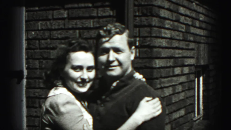 The couple turn toward the camera and smile while embracing against a brick wall in the supplied 1946 Pennsylvania setting.