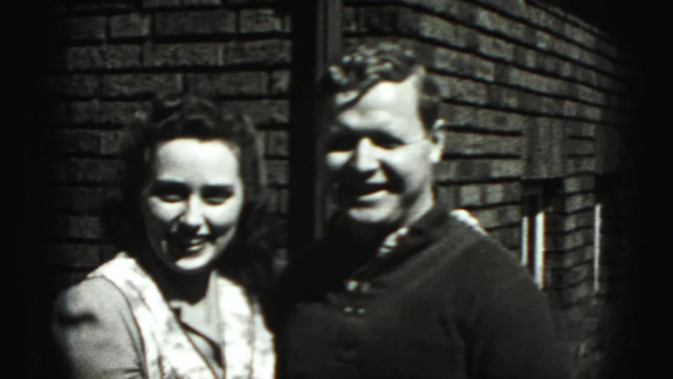 A man and woman smile toward the camera with arms around each other beside a brick wall in this 1940s home-movie frame.