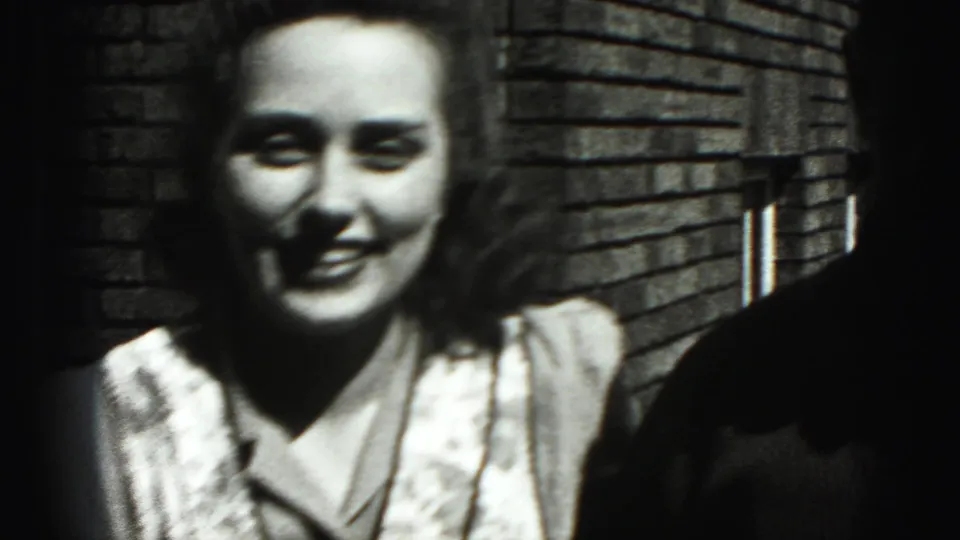 Close 8mm frame of the woman smiling near the camera, with the man partly visible at the edge and the brick wall behind.