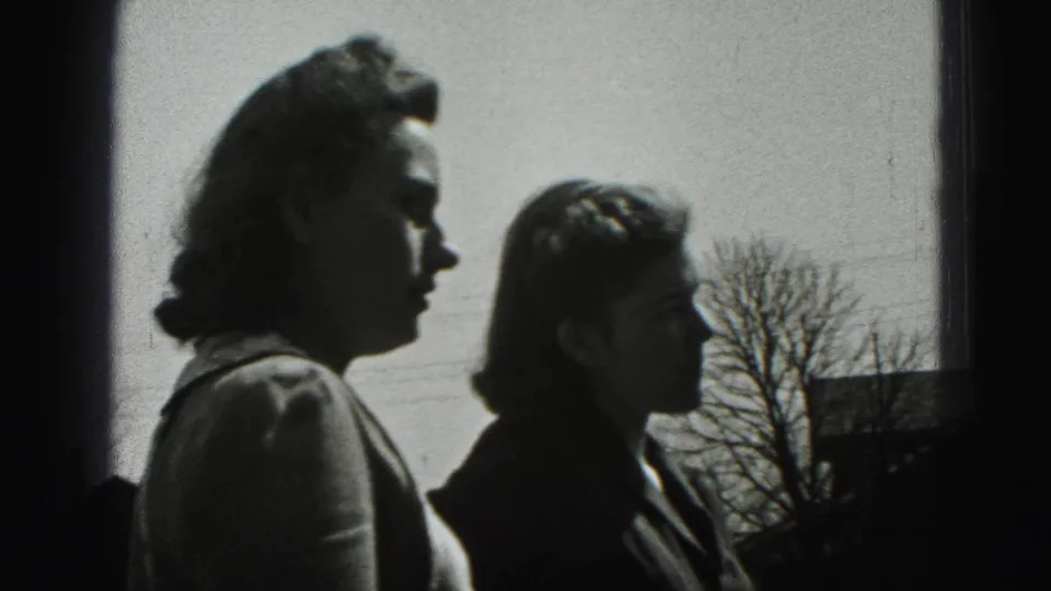 Two women stand in profile outdoors in New Cumberland, with a bare tree and distant rooftops visible behind them.