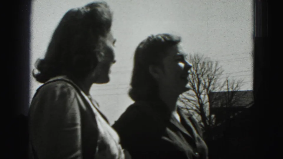 The women appear in side view in the 1946 home-movie scene, framed against a bright sky, bare tree, and nearby roofline.