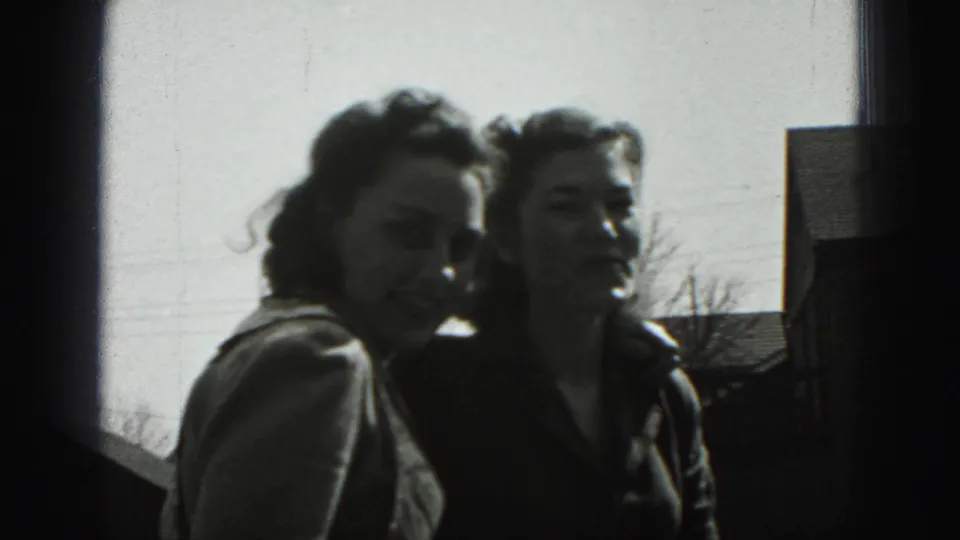 In New Cumberland, two young women lean together and look toward the camera, with a bare tree and house roof behind them.