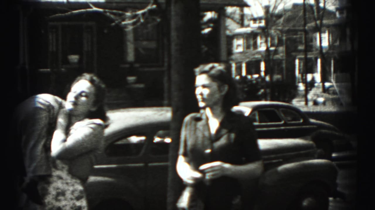 Woman carries a small child while another woman stands near a parked sedan.