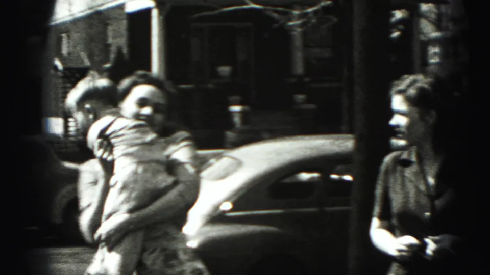 Two women and a small child move along a New Cumberland sidewalk with a dark 1940s car behind them.