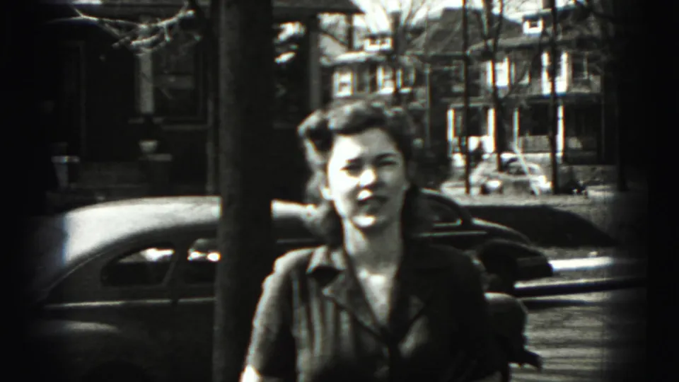 A young woman faces the camera on a New Cumberland residential street, with a parked sedan and houses in the background.