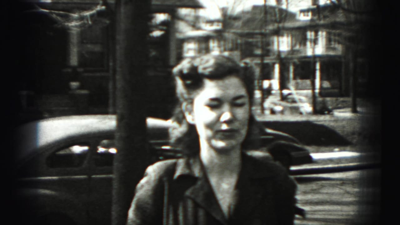 Woman in a dark dress standing on a residential street with parked cars behind her.