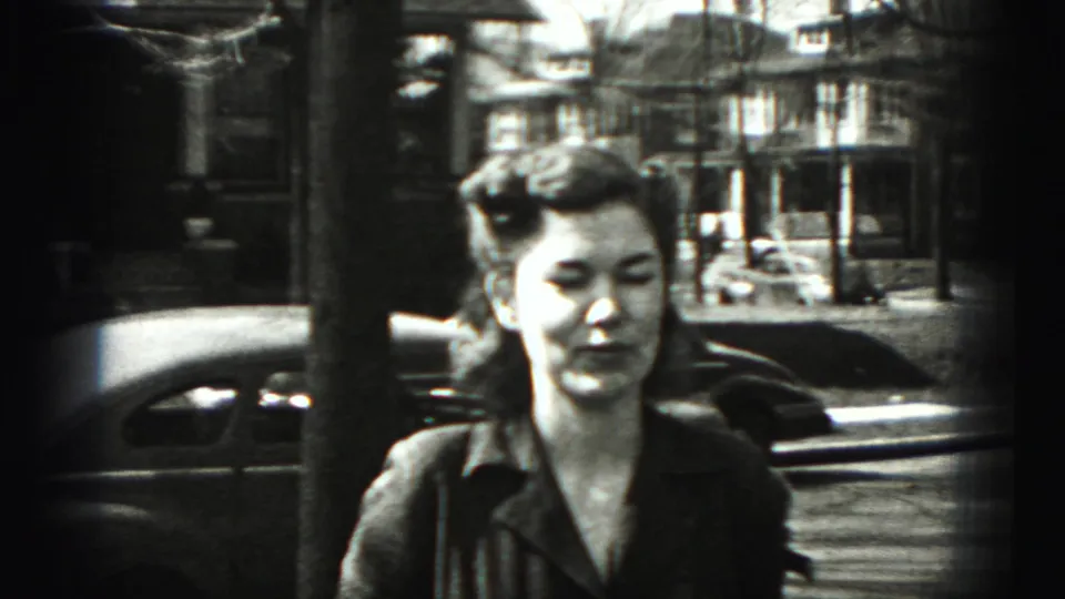 In 1946 New Cumberland, Pennsylvania footage, a woman stands near a tree-lined residential street with rounded cars in the background.