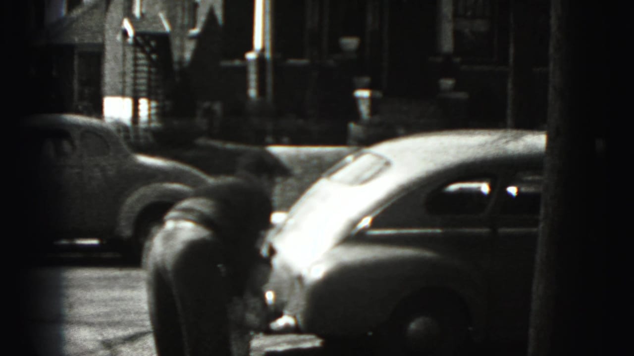 The camera shifts away from the woman to a New Cumberland residential street view with rounded 1940s cars and house fronts.