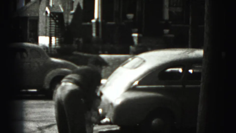 The camera shifts away from the woman to a New Cumberland residential street view with rounded 1940s cars and house fronts.