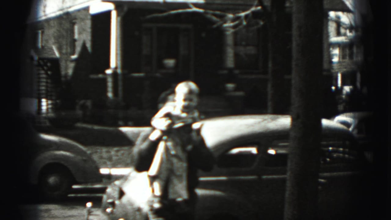 An adult appears near a parked car on the New Cumberland street, holding a small figure, with houses and trees behind.