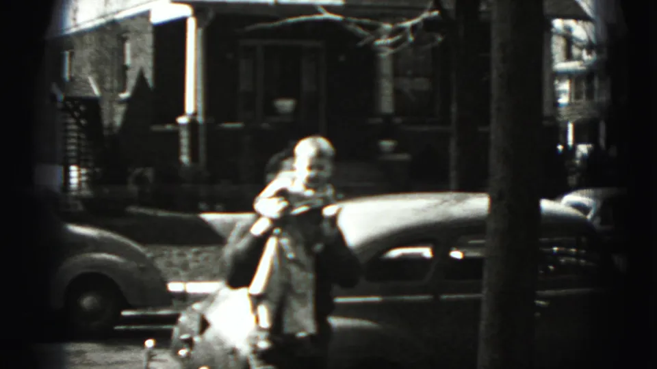 An adult appears near a parked car on the New Cumberland street, holding a small figure, with houses and trees behind.