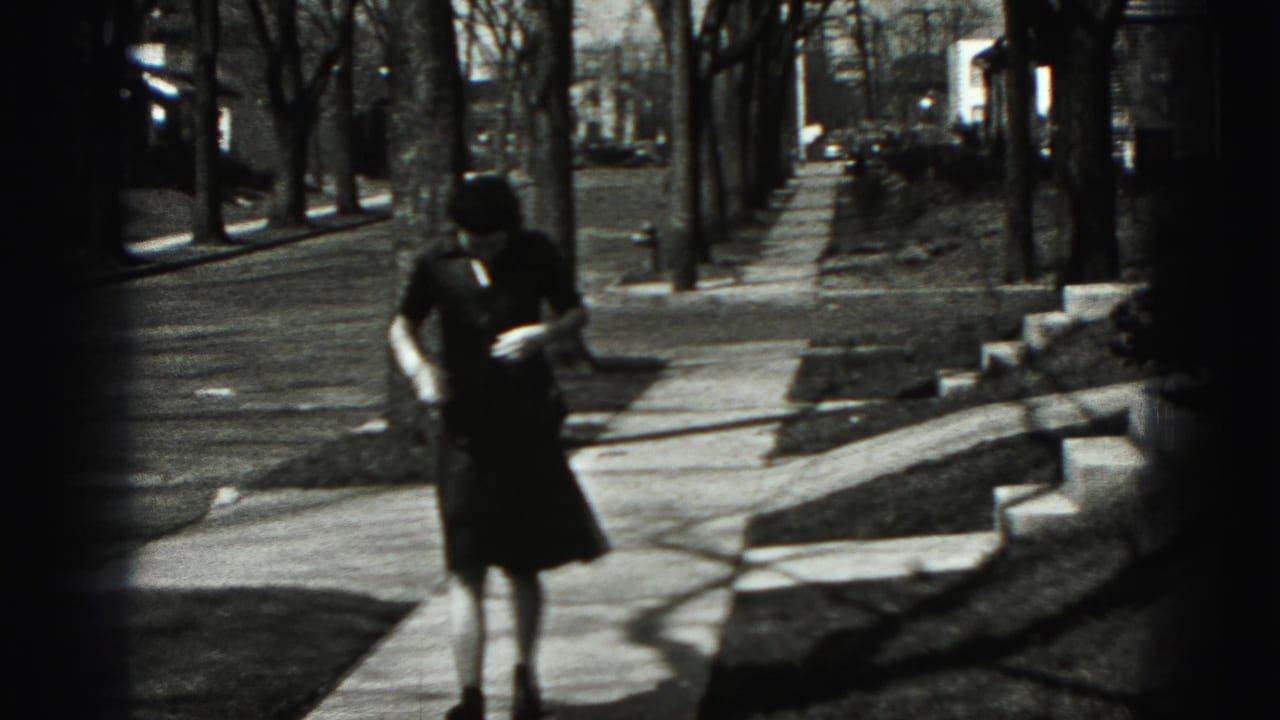 The woman continues along the New Cumberland sidewalk, framed by bare trees, lawn edges, and residential buildings.