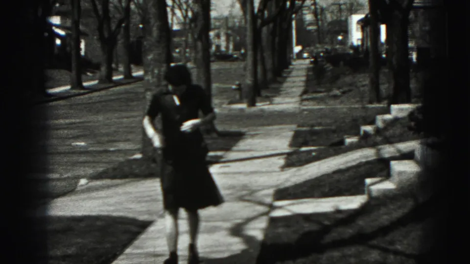 The woman continues along the New Cumberland sidewalk, framed by bare trees, lawn edges, and residential buildings.
