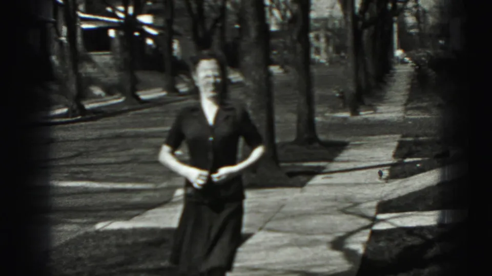 The young woman faces the camera and appears to button or adjust her blouse during the 1946 home-movie sidewalk shot.