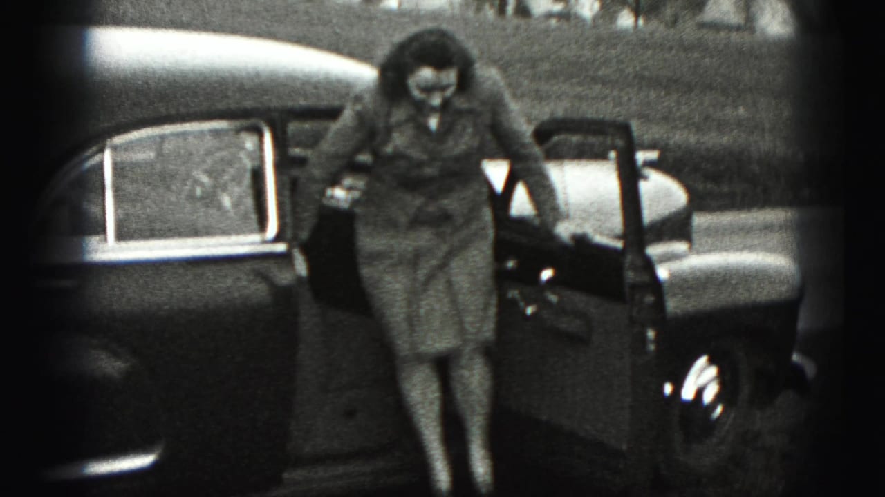 In 1946 New Cumberland, Pennsylvania, a woman in a tailored suit steps down from a rounded sedan beside a grassy roadside.