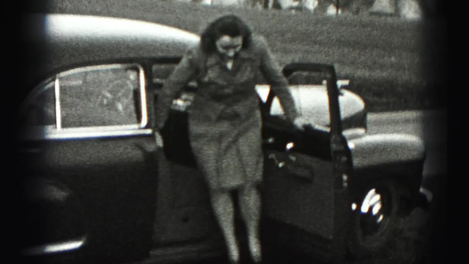 In 1946 New Cumberland, Pennsylvania, a woman in a tailored suit steps down from a rounded sedan beside a grassy roadside.