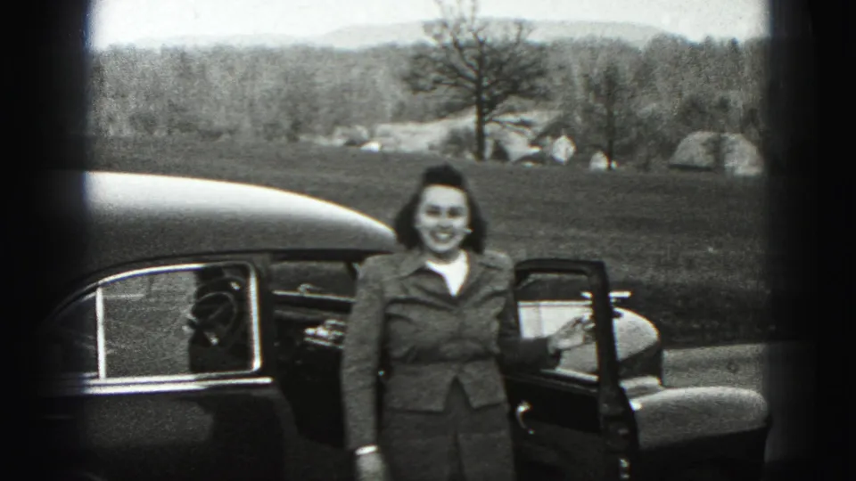 The woman stands smiling beside the open sedan door, with grass, trees, rocks, and distant hills behind her.