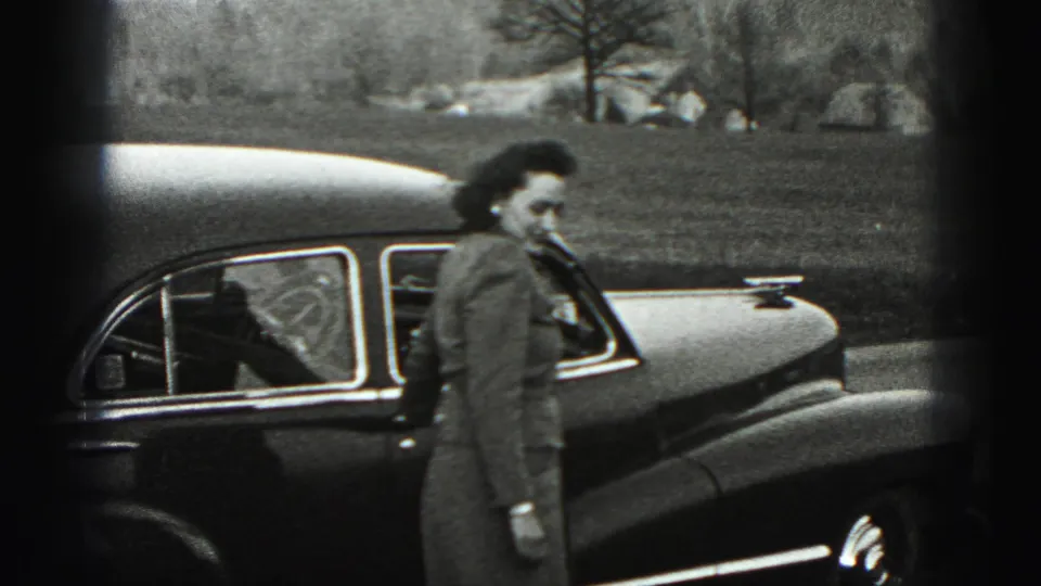 The woman walks along the side of the rounded sedan, with open grass, bare trees, and low hills in the background.