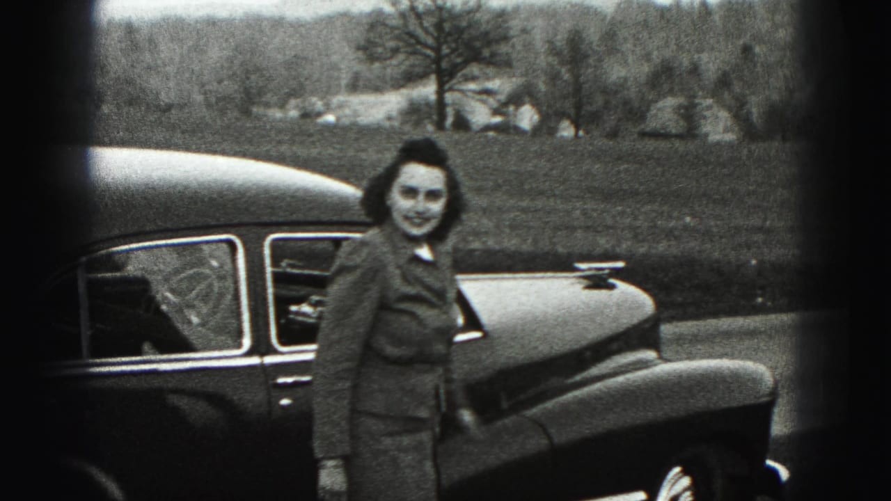 In 1946 New Cumberland, Pennsylvania, a woman in a tailored suit faces the camera beside a rounded dark car in an outdoor setting.